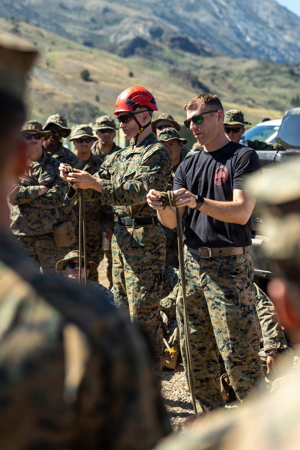 Two instructors are conducting an outdoor military training demonstration involving rope or cordage techniques for a group of assembled personnel. One instructor wears a red climbing helmet and MARPAT camouflage uniform, while the other wears a black t-shirt with a unit insignia and MARPAT trousers. A large group of uniformed military personnel observes in the background against a mountainous terrain.