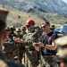 Two instructors are conducting an outdoor military training demonstration involving rope or cordage techniques for a group of assembled personnel. One instructor wears a red climbing helmet and MARPAT camouflage uniform, while the other wears a black t-shirt with a unit insignia and MARPAT trousers. A large group of uniformed military personnel observes in the background against a mountainous terrain.