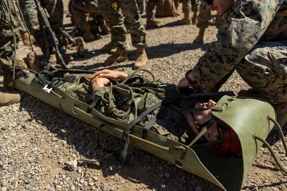 Military personnel in MARPAT camouflage uniforms are conducting a casualty evacuation (CASEVAC) training exercise in an arid, gravel terrain. A simulated casualty is secured in an olive-drab Sked stretcher (flexible rescue litter) on the ground, with a fellow service member kneeling beside them providing assistance. A group of additional uniformed personnel observe the exercise in the background.