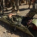 Military personnel in MARPAT camouflage uniforms are conducting a casualty evacuation (CASEVAC) training exercise in an arid, gravel terrain. A simulated casualty is secured in an olive-drab Sked stretcher (flexible rescue litter) on the ground, with a fellow service member kneeling beside them providing assistance. A group of additional uniformed personnel observe the exercise in the background.