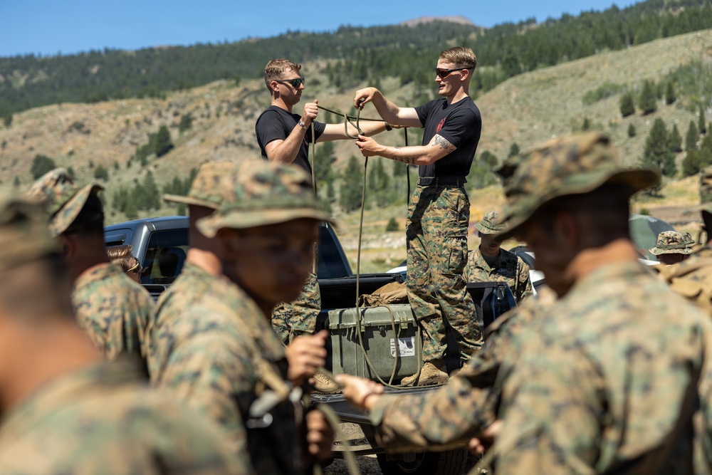 A group of military personnel in camouflage uniforms are gathered outdoors in a mountainous, forested setting. Two individuals standing on the bed of a pickup truck are demonstrating rope or cord handling techniques to the assembled group. The scene appears to be a field training exercise or instruction session.
