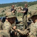 A group of military personnel in camouflage uniforms are gathered outdoors in a mountainous, forested setting. Two individuals standing on the bed of a pickup truck are demonstrating rope or cord handling techniques to the assembled group. The scene appears to be a field training exercise or instruction session.