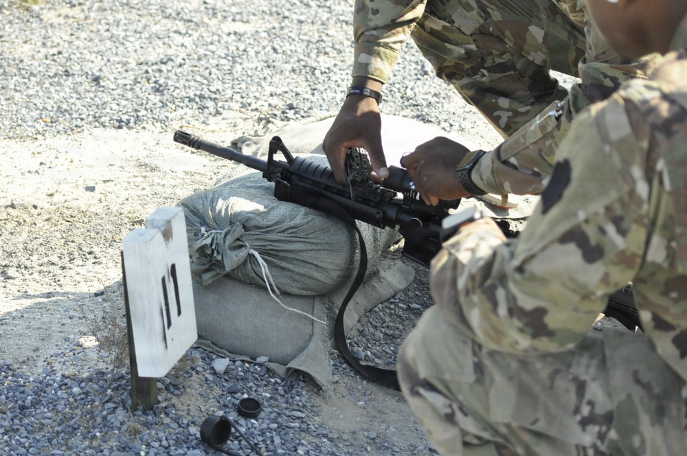 Two military personnel in MultiCam/OCP camouflage uniforms are handling an M4/M16-style carbine rifle at what appears to be a firing range. The weapon is resting on sandbags at a numbered shooting position marked '11'. The setting is an outdoor gravel range during daylight hours.
