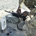 Two military personnel in MultiCam/OCP camouflage uniforms are handling an M4/M16-style carbine rifle at what appears to be a firing range. The weapon is resting on sandbags at a numbered shooting position marked '11'. The setting is an outdoor gravel range during daylight hours.