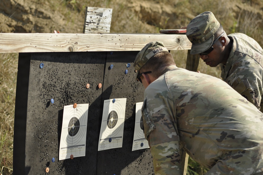 Two military personnel in MultiCam/OCP camouflage uniforms are examining paper shooting targets on a range backstop. They appear to be scoring or assessing shot placement on standard bullseye-style qualification targets. The setting is an outdoor firing range with a dirt berm backstop visible in the background.