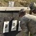 Two military personnel in MultiCam/OCP camouflage uniforms are examining paper shooting targets on a range backstop. They appear to be scoring or assessing shot placement on standard bullseye-style qualification targets. The setting is an outdoor firing range with a dirt berm backstop visible in the background.