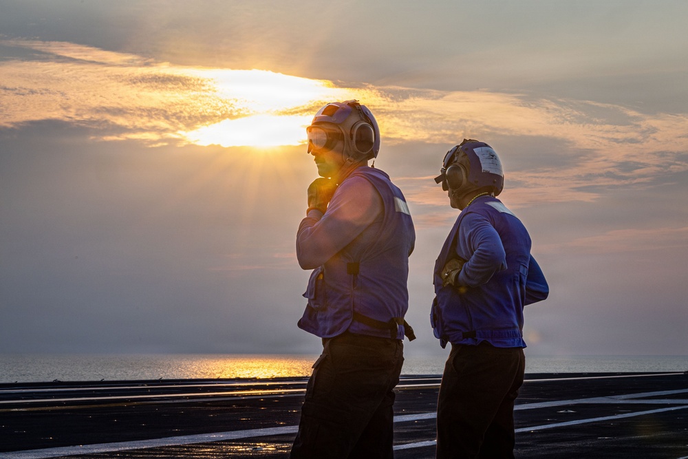 Two personnel stand on what appears to be an aircraft carrier flight deck at sunrise or sunset, silhouetted against a dramatic sky with the sun breaking through clouds over open water. Both individuals are wearing flight deck safety gear including cranial helmets with ear protection and colored safety vests. The wet, dark surface of the flight deck is visible in the foreground.