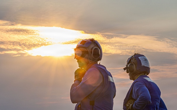 Two personnel stand on what appears to be an aircraft carrier flight deck at sunrise or sunset, silhouetted against a dramatic sky with the sun breaking through clouds over open water. Both individuals are wearing flight deck safety gear including cranial helmets with ear protection and colored safety vests. The wet, dark surface of the flight deck is visible in the foreground.