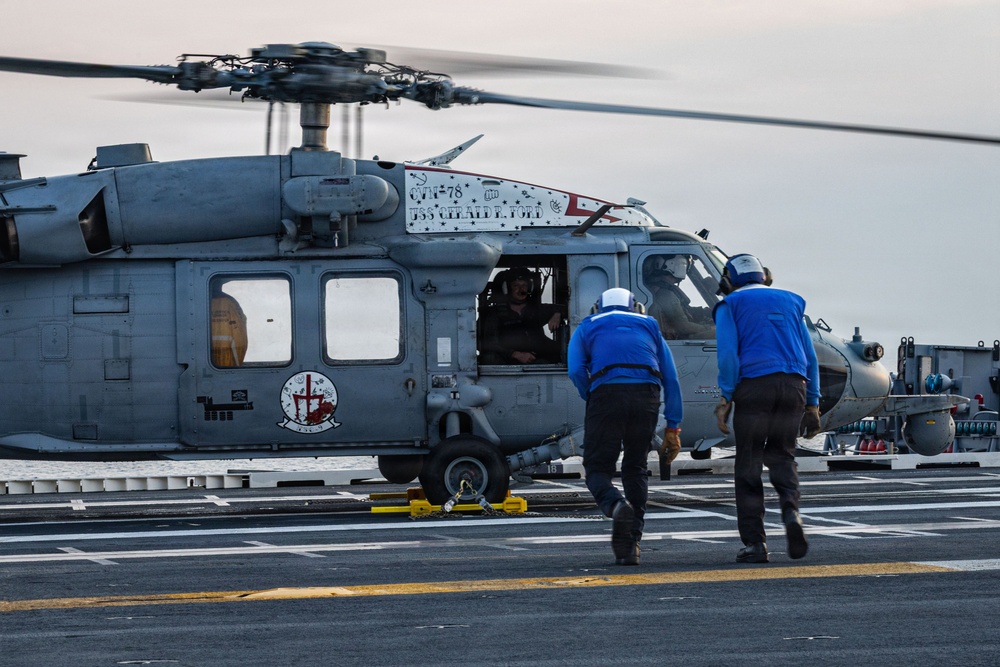 A military helicopter, marked with 'USS Gerald R. Ford' and 'CTF-78' designations, is on the flight deck of a naval aircraft carrier. Two deck crew members in blue uniforms are approaching the helicopter, which has its rotors spinning and a pilot visible through the cockpit window. Yellow wheel chocks are visible securing the landing gear to the deck.