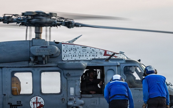 A military helicopter, marked with 'USS Gerald R. Ford' and 'CTF-78' designations, is on the flight deck of a naval aircraft carrier. Two deck crew members in blue uniforms are approaching the helicopter, which has its rotors spinning and a pilot visible through the cockpit window. Yellow wheel chocks are visible securing the landing gear to the deck.