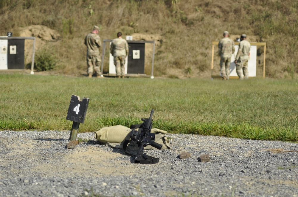 A military shooting range is depicted with personnel in camouflage uniforms inspecting targets downrange. In the foreground, an assault rifle (appears to be an M4/M16-style carbine) rests on a sandbag next to a lane marker labeled '4'. Multiple target stands are visible in the background along a berm.