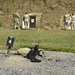 A military shooting range is depicted with personnel in camouflage uniforms inspecting targets downrange. In the foreground, an assault rifle (appears to be an M4/M16-style carbine) rests on a sandbag next to a lane marker labeled '4'. Multiple target stands are visible in the background along a berm.