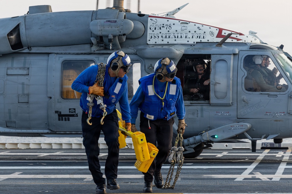 Two Navy deck crew members in blue jerseys are carrying tie-down chains and a yellow chock device away from a military helicopter on the flight deck of USS Gerald R. Ford (CVN-78). The helicopter, an MH-60 Seahawk, displays the ship's name and hull number on a placard. At least two aircrew members are visible inside the helicopter cockpit.