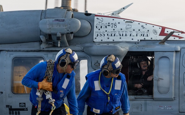Two Navy deck crew members in blue jerseys are carrying tie-down chains and a yellow chock device away from a military helicopter on the flight deck of USS Gerald R. Ford (CVN-78). The helicopter, an MH-60 Seahawk, displays the ship's name and hull number on a placard. At least two aircrew members are visible inside the helicopter cockpit.