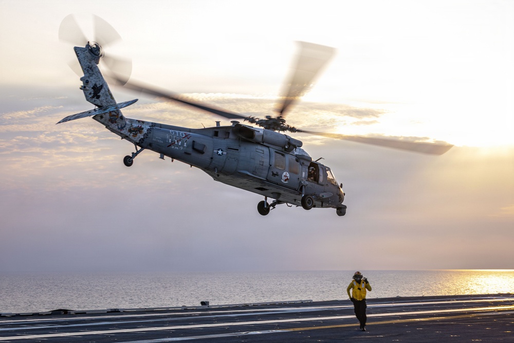 A U.S. Navy MH-60 Seahawk helicopter is captured mid-hover or during takeoff/landing operations on the flight deck of an aircraft carrier at sunset or sunrise. A flight deck crew member in a yellow jersey is visible on the deck, appearing to communicate via radio or headset while observing the helicopter. The scene is set over open ocean with warm golden light reflecting off calm waters in the background.