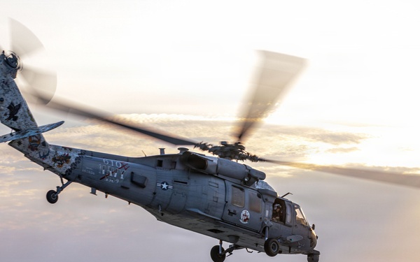 A U.S. Navy MH-60 Seahawk helicopter is captured mid-hover or during takeoff/landing operations on the flight deck of an aircraft carrier at sunset or sunrise. A flight deck crew member in a yellow jersey is visible on the deck, appearing to communicate via radio or headset while observing the helicopter. The scene is set over open ocean with warm golden light reflecting off calm waters in the background.
