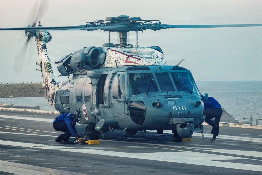 A military helicopter numbered '610' is parked on what appears to be a naval vessel's flight deck, with the ocean visible in the background. The helicopter features distinctive decorative artwork on its tail section and top, suggesting it may be a special commemorative or squadron-marked aircraft. Ground crew members dressed in blue uniforms are performing maintenance or securing operations around the aircraft.