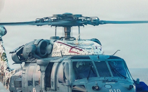 A military helicopter numbered '610' is parked on what appears to be a naval vessel's flight deck, with the ocean visible in the background. The helicopter features distinctive decorative artwork on its tail section and top, suggesting it may be a special commemorative or squadron-marked aircraft. Ground crew members dressed in blue uniforms are performing maintenance or securing operations around the aircraft.