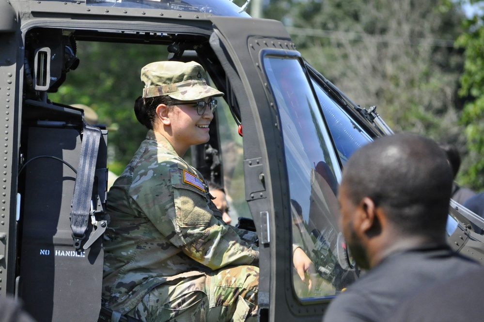 A smiling U.S. Army soldier in OCP (Operational Camouflage Pattern) uniform is seated inside a military helicopter, likely during a public outreach or demonstration event. She is wearing glasses and a patrol cap with a U.S. flag patch visible on her shoulder. A civilian individual is visible in the foreground, suggesting an interaction or tour scenario.