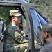 A smiling U.S. Army soldier in OCP (Operational Camouflage Pattern) uniform is seated inside a military helicopter, likely during a public outreach or demonstration event. She is wearing glasses and a patrol cap with a U.S. flag patch visible on her shoulder. A civilian individual is visible in the foreground, suggesting an interaction or tour scenario.