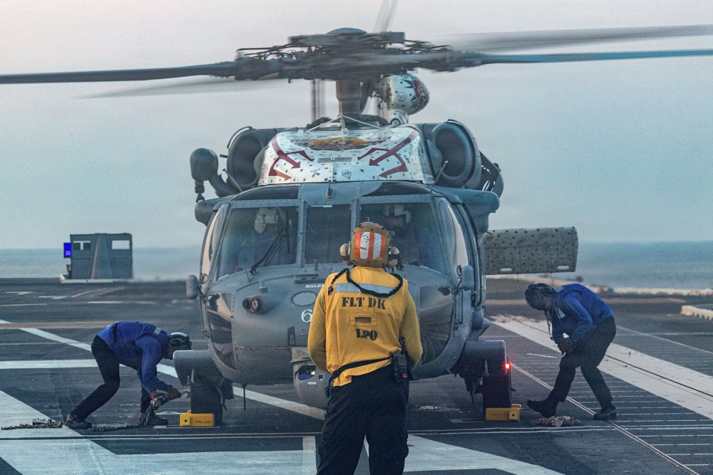 A military helicopter, likely an MH-60 Seahawk, is on the flight deck of a naval vessel with rotor blades spinning. Three crew members are visible: one in a yellow jacket labeled 'FLT DK LPO' standing centrally, and two others in blue working at the helicopter's landing gear area. The scene appears to be taking place at dusk or dawn in overcast conditions.