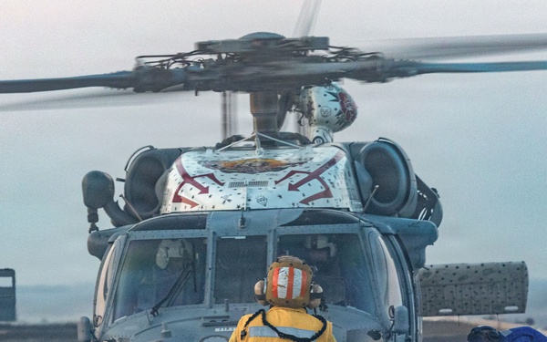 A military helicopter, likely an MH-60 Seahawk, is on the flight deck of a naval vessel with rotor blades spinning. Three crew members are visible: one in a yellow jacket labeled 'FLT DK LPO' standing centrally, and two others in blue working at the helicopter's landing gear area. The scene appears to be taking place at dusk or dawn in overcast conditions.