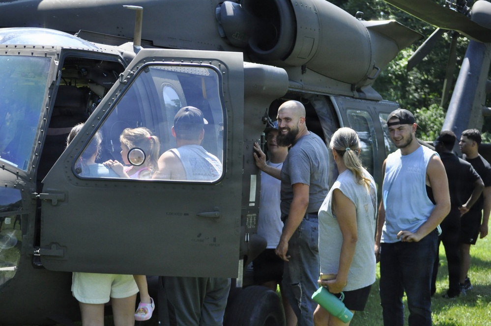 A group of civilians is gathered around a military helicopter, likely a UH-60 Black Hawk, during what appears to be a public open house or air show event. Adults and children are examining the aircraft up close, with some looking into the cockpit and others standing near the cabin door. The setting is an outdoor grassy area with trees visible in the background.