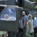 A group of civilians is gathered around a military helicopter, likely a UH-60 Black Hawk, during what appears to be a public open house or air show event. Adults and children are examining the aircraft up close, with some looking into the cockpit and others standing near the cabin door. The setting is an outdoor grassy area with trees visible in the background.