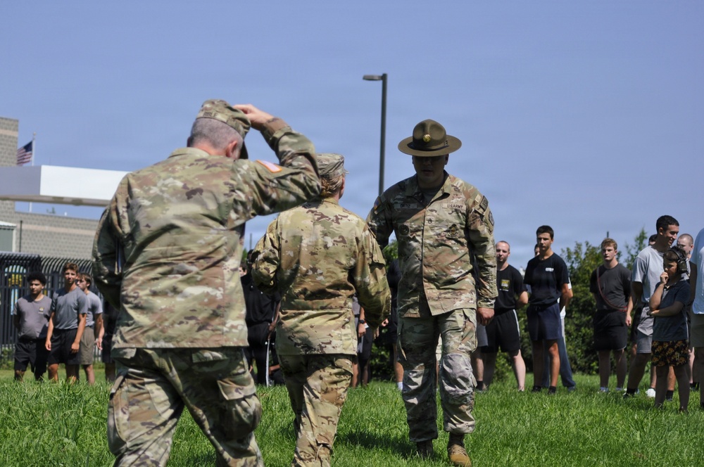 An outdoor military scene showing uniformed U.S. Army personnel interacting on a grass field. One soldier is rendering a salute while a drill sergeant wearing a distinctive campaign (Smokey Bear) hat faces them. A group of civilians or recruits in civilian athletic wear observe from the background.