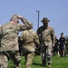 An outdoor military scene showing uniformed U.S. Army personnel interacting on a grass field. One soldier is rendering a salute while a drill sergeant wearing a distinctive campaign (Smokey Bear) hat faces them. A group of civilians or recruits in civilian athletic wear observe from the background.