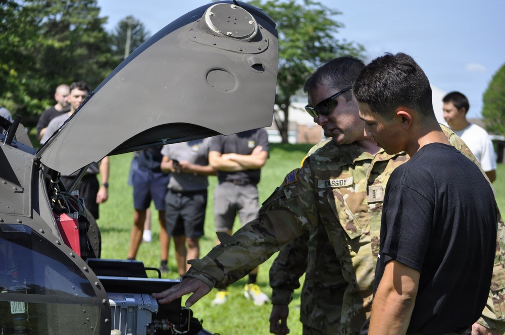A U.S. Army soldier in multicam camouflage uniform, identified by the name tape 'CASSIDY', is demonstrating or explaining the internal components of a military aircraft (likely an Apache helicopter) to a group of individuals in civilian and military attire. The scene appears to be an outdoor demonstration or educational event on a military installation. Several observers stand in the background watching the presentation.