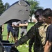A U.S. Army soldier in multicam camouflage uniform, identified by the name tape 'CASSIDY', is demonstrating or explaining the internal components of a military aircraft (likely an Apache helicopter) to a group of individuals in civilian and military attire. The scene appears to be an outdoor demonstration or educational event on a military installation. Several observers stand in the background watching the presentation.