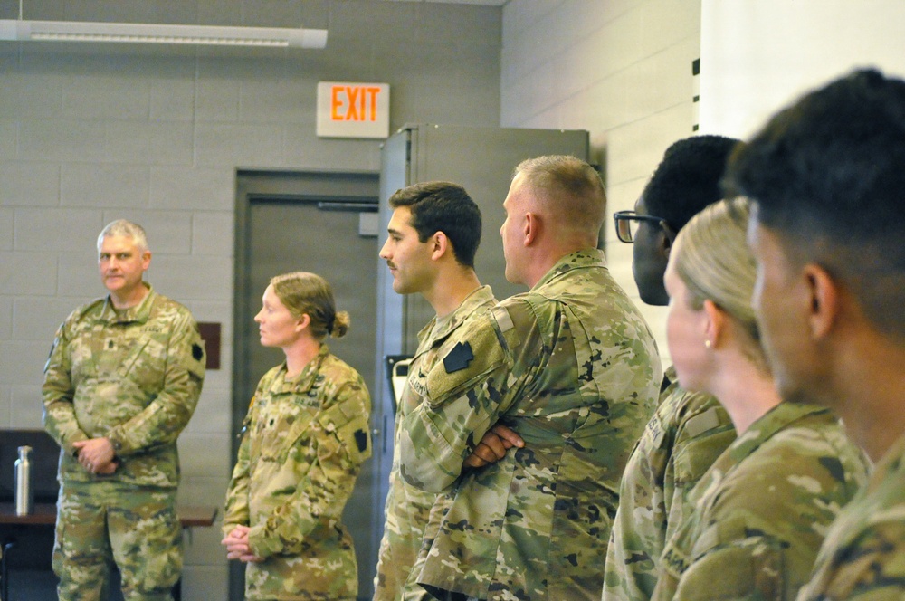 A group of military personnel in US Army OCP (Operational Camouflage Pattern) uniforms are standing in a line inside what appears to be a military facility or briefing room. They appear to be attentively listening to a presentation or briefing, facing the same direction. The group is mixed in gender and ethnicity, suggesting a diverse unit assembly.