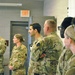 A group of military personnel in US Army OCP (Operational Camouflage Pattern) uniforms are standing in a line inside what appears to be a military facility or briefing room. They appear to be attentively listening to a presentation or briefing, facing the same direction. The group is mixed in gender and ethnicity, suggesting a diverse unit assembly.