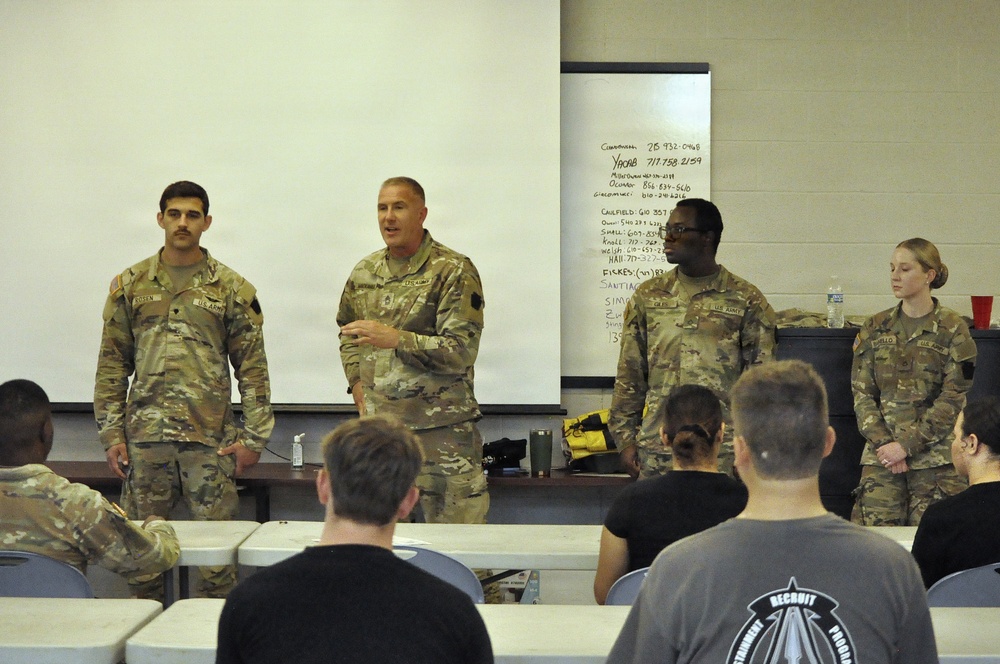 Four U.S. Army soldiers in OCP (Operational Camouflage Pattern) uniforms stand at the front of a classroom-style briefing room, with one soldier appearing to address an audience seated in rows. The setting appears to be a military training or administrative briefing environment, with a whiteboard behind them containing handwritten notes and phone numbers. Civilian or military personnel in civilian attire are seated facing the presenters.