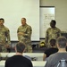 Four U.S. Army soldiers in OCP (Operational Camouflage Pattern) uniforms stand at the front of a classroom-style briefing room, with one soldier appearing to address an audience seated in rows. The setting appears to be a military training or administrative briefing environment, with a whiteboard behind them containing handwritten notes and phone numbers. Civilian or military personnel in civilian attire are seated facing the presenters.