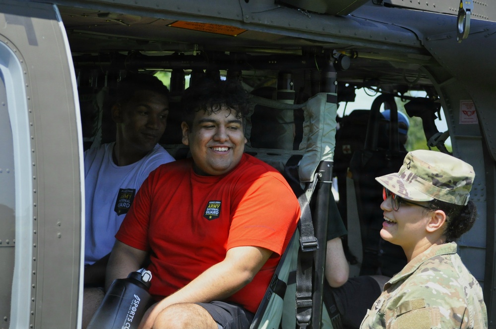 A soldier in Army Combat Uniform (ACU) interacts with two young civilians seated inside a military helicopter, likely during a public outreach or recruitment event. The civilians are wearing Army National Guard branded t-shirts, suggesting participation in a youth program or community engagement activity. All individuals appear to be smiling and engaged in friendly conversation.
