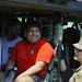 A soldier in Army Combat Uniform (ACU) interacts with two young civilians seated inside a military helicopter, likely during a public outreach or recruitment event. The civilians are wearing Army National Guard branded t-shirts, suggesting participation in a youth program or community engagement activity. All individuals appear to be smiling and engaged in friendly conversation.