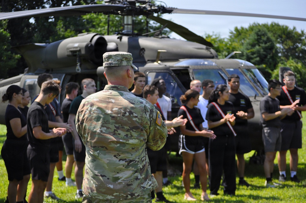 A U.S. Army soldier in OCP (Operational Camouflage Pattern) uniform, identified by the name tape 'KRENN', addresses a group of young civilians standing in front of two military UH-60 Black Hawk helicopters on a grassy field. The setting appears to be a recruitment or outreach event, possibly a JROTC or Army recruiting demonstration. The group of young people, dressed in casual athletic wear, are attentively listening to the briefing.