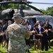 A U.S. Army soldier in OCP (Operational Camouflage Pattern) uniform, identified by the name tape 'KRENN', addresses a group of young civilians standing in front of two military UH-60 Black Hawk helicopters on a grassy field. The setting appears to be a recruitment or outreach event, possibly a JROTC or Army recruiting demonstration. The group of young people, dressed in casual athletic wear, are attentively listening to the briefing.