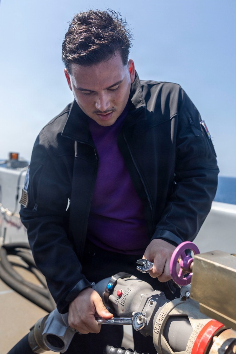 A young man wearing a black jacket over a purple shirt is working on mechanical equipment, likely a valve or pipe fitting assembly, on what appears to be the deck of a naval or maritime vessel. He is using wrenches to tighten or adjust components on the equipment. The background shows open water and a clear sky, confirming an outdoor maritime setting.