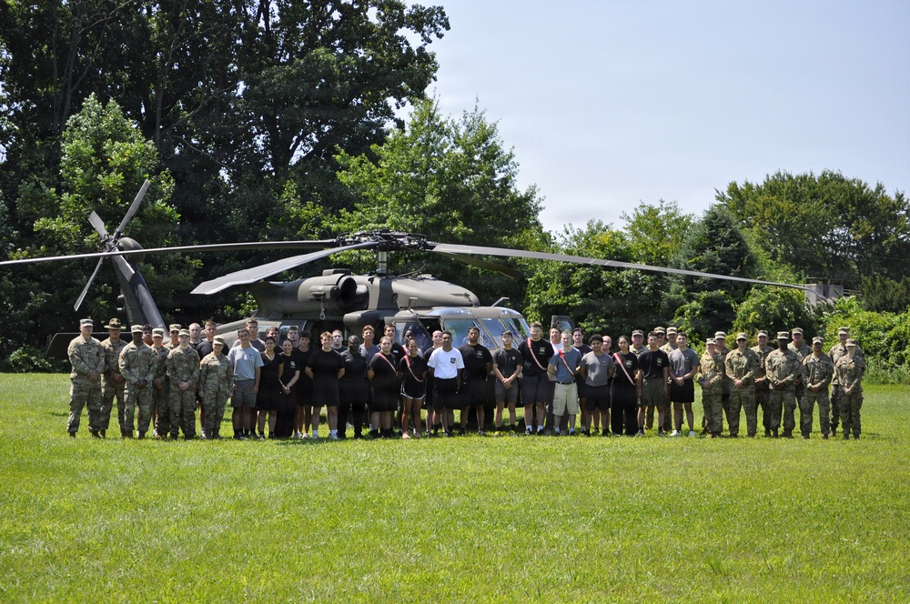 A large group of military personnel and civilians pose for a group photograph in front of a UH-60 Black Hawk helicopter on a grassy field. The group includes uniformed soldiers in Army Combat Uniforms (ACU/OCP pattern) as well as individuals in civilian or PT (physical training) attire, suggesting a mixed military and possibly ROTC or recruiting event. The setting appears to be an outdoor field surrounded by trees on a clear summer day.