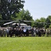 A large group of military personnel and civilians pose for a group photograph in front of a UH-60 Black Hawk helicopter on a grassy field. The group includes uniformed soldiers in Army Combat Uniforms (ACU/OCP pattern) as well as individuals in civilian or PT (physical training) attire, suggesting a mixed military and possibly ROTC or recruiting event. The setting appears to be an outdoor field surrounded by trees on a clear summer day.