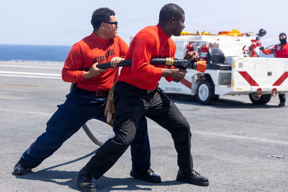 Two personnel wearing red shirts labeled 'CRASH' and black pants are conducting a firefighting hose drill on what appears to be a naval vessel's flight deck. They are bracing against the pressure of a fire hose with a nozzle attachment, practicing firefighting techniques. A white crash/fire/rescue vehicle with red markings is visible in the background, along with a third person in a black balaclava.