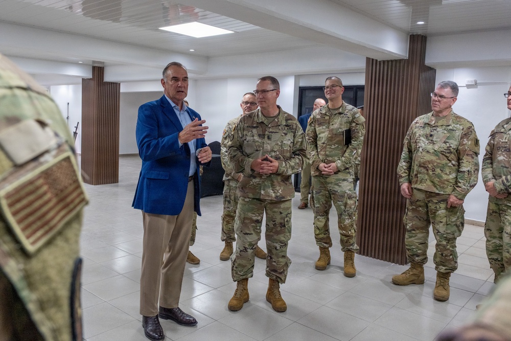 A civilian in a blue blazer and khaki pants appears to be briefing or addressing a group of U.S. Army soldiers in OCP (Operational Camouflage Pattern) uniforms inside a modern, well-lit facility. The soldiers are standing attentively in a semi-circle, listening to the speaker. The setting appears to be an official meeting or visit at a military or government installation.