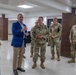 A civilian in a blue blazer and khaki pants appears to be briefing or addressing a group of U.S. Army soldiers in OCP (Operational Camouflage Pattern) uniforms inside a modern, well-lit facility. The soldiers are standing attentively in a semi-circle, listening to the speaker. The setting appears to be an official meeting or visit at a military or government installation.