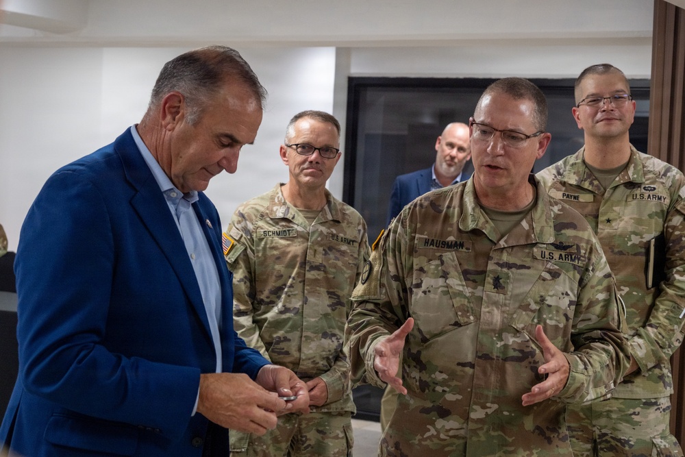 A group of U.S. Army personnel in OCP (Operational Camouflage Pattern) uniforms are engaged in a meeting or briefing with a civilian man in a blue blazer. The central Army officer, identified by his name tape as 'HAUSMAN,' appears to be speaking and gesturing while the civilian examines a small object. Two other uniformed soldiers, identified as 'SCHMIDT' and 'PAYNE,' are visible in the background along with another civilian.