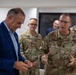 A group of U.S. Army personnel in OCP (Operational Camouflage Pattern) uniforms are engaged in a meeting or briefing with a civilian man in a blue blazer. The central Army officer, identified by his name tape as 'HAUSMAN,' appears to be speaking and gesturing while the civilian examines a small object. Two other uniformed soldiers, identified as 'SCHMIDT' and 'PAYNE,' are visible in the background along with another civilian.