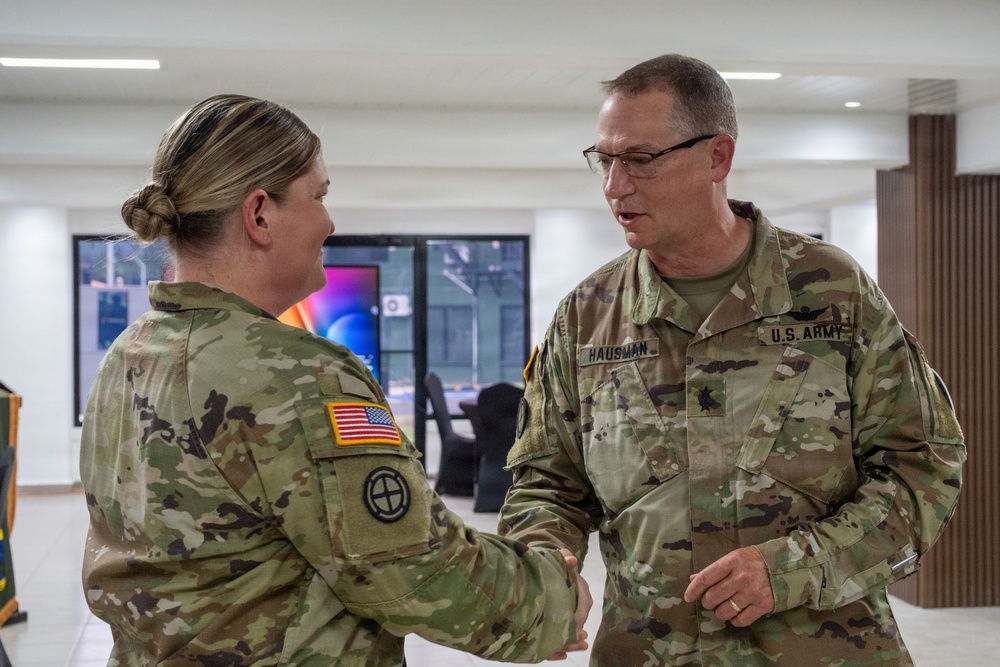 Two U.S. Army soldiers in OCP (Operational Camouflage Pattern) uniforms are shaking hands in what appears to be a modern indoor facility. The male soldier on the right has a visible name tape reading 'HAUSMAN' and wears a U.S. Army tape, along with rank insignia consistent with a Major (oak leaf cluster). The interaction appears to be a formal or ceremonial exchange.