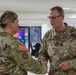 Two U.S. Army soldiers in OCP (Operational Camouflage Pattern) uniforms are shaking hands in what appears to be a modern indoor facility. The male soldier on the right has a visible name tape reading 'HAUSMAN' and wears a U.S. Army tape, along with rank insignia consistent with a Major (oak leaf cluster). The interaction appears to be a formal or ceremonial exchange.